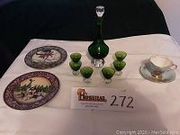 Green cordial decanter and six matching cordial glasses with two decorative plates and one bone china cup and saucer set on white cloth.