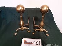 Front view of two brass andirons showing ball finials and claw feet on a dark fabric background.