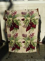 Full view of one barkcloth drapery panel displayed on a wooden fabric rack in daylight, showing botanical print of green and burgundy leaves on beige background.