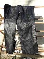 Black Harley Davidson leather motorcycle chaps, folded over a wooden railing with clear embossed logo visible