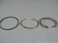 Three silver bracelets lying flat on a white surface; shows the variety of designs: a plain bangle, an irregular shaped cuff, and a twisted open cuff.