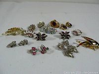 Photo showing an overview of assorted vintage costume jewelry pieces including brooches and earrings arranged on a white surface.