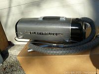 Side view of vintage Electrolux vacuum with braided hose and metal wand attachment, placed on cardboard.