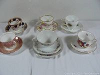 Photo showing six antique vintage assorted porcelain teacups with matching saucers arranged on a flat surface, featuring floral and decorative patterns with gold trim on some pieces.