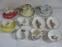 Front angled view of nine assorted vintage porcelain tea and tableware pieces including cups, saucers, plates, and creamer, showing floral designs and gold accents.