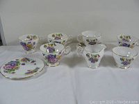 Six vintage white china tea cups and one matching saucer with floral pattern and gold trim arranged on white cloth.