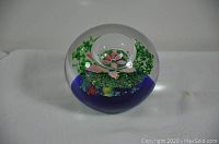 Front view of clear spherical glass paperweight with pink flower and green leaves inside, sitting on a blue base.
