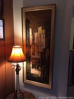 Full view of the framed vertical oil on board painting hanging on wall adjacent to a lamp.