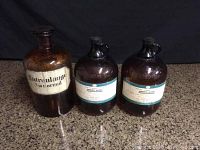 Three vintage large brown glass medicine bottles on granite counter, two labeled Isopropyl Alcohol and one labeled Natronlauge 1/10 Normal, showing overall lot.