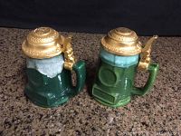Two vintage ceramic tankards with green and white coloration and brass lids, placed together on granite surface