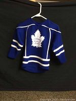 Full front view of blue Toronto Maple Leafs kids hockey jersey with white stripes and large white maple leaf logo on front.