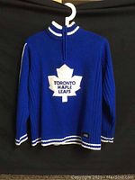Full view of blue knitted wool sweater with quarter-zip collar and Toronto Maple Leafs white maple leaf logo patch on chest, showing striped trim at collar, cuffs, and hem.