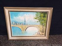 Full view of framed oil painting showing stone arch bridge, river, cityscape and trees.