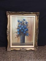 Front view of the framed oil painting showing a bouquet of blue flowers in gray vase against a warm toned background.