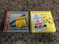 Photo of two vintage 8mm film reels on a granite surface: 'Adventures in Disneyland, Frontierland' and 'The Three Stooges in Eye Doctors'. Both are in original cardboard sleeves.