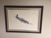 Framed numbered print of a Spitfire fighter plane by John Moutray shown hanging on a wall.
