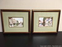 Two framed landscape prints with wood frames and green double mattes, showing village scenes