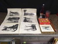 Six black and white prints laid flat with additional framed print, lava lamp, Coca Cola puzzle box, glass cup, and wooden item visible