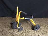 Full side view of yellow RABO kids tricycle with black plastic seat and three solid rubber wheels on a brown speckled floor.