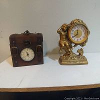 Wooden vintage style clock with round white dial and black numbers beside a decorative gold-tone clock with a figurine of a child holding the clock face. Both clocks shown on white surface against blank wall.