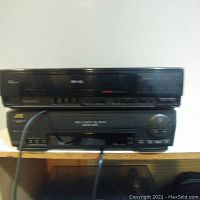 Photo of two vintage black VCR players stacked on shelf. Top unit is General Electric, bottom unit is JVC. Both have front-facing control buttons and tape slots. A cable hangs from the front of the JVC unit.