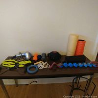 Full display of exercise equipment including massager, resistance bands, massage balls, roller, weighted bag, jump rope, and loop band on a wooden table against wall.