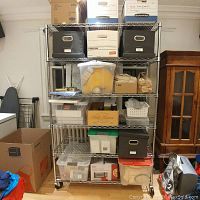 Photo showing the stainless steel wire storage rack on casters loaded with various boxes and containers. Walls, ironing board, wooden cabinet, and other objects visible around.
