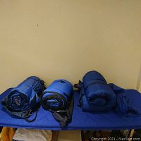 Three blue sleeping bags rolled up on a blue surface, showing the size and condition of each sleeping bag in their stuff sacks.