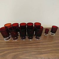 12 red glass drinking glasses arranged on a wooden surface including 10 large and 2 smaller ones with clear pedestal bases
