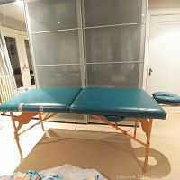 Full view of teal padded massage table with foldable wooden legs and attached headrest pillow.