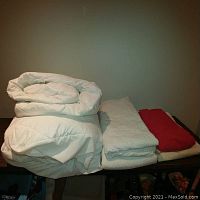 Photo showing folded white down mattress topper, two white mattress covers, and folded white and red linens on a flat surface.