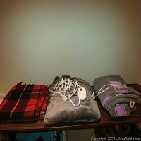 Four blankets on a wooden table: two gray electric blankets with cords and controls, and two folded wool throw blankets in plaid patterns, one red and black, the other red with multicolor plaid.