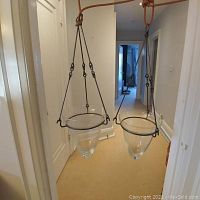 Two hanging glass vases with black metal frames suspended from an orange rope in a hallway.