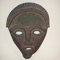 Front view of a hand-carved wooden Indonesian mask featuring muted earth tone colors, geometric pattern headdress and stylized human face with open eyes and mouth.