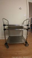 Front view of the stainless steel trolley showing three shelves, side handles, and swivel wheels.