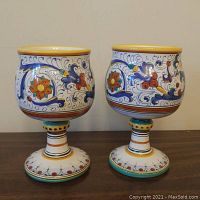 Front view of two hand-painted Italian porcelain goblets with multi-color floral and scroll patterns on pedestal bases.