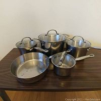Five cookware pieces displayed on tabletop: three lidded pots and one lidded saucepan at back, one lidless sauté pan at front.