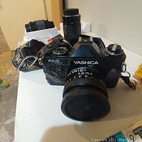 Black Yashica FX-D Quartz camera with attached Yashica 50mm f/2 lens and patterned fabric strap, placed on white surface.