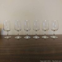 Front view showing 6 of the wine glasses arranged in a row on a table against a white wall.