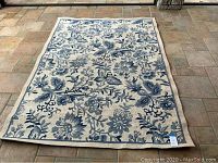 Full view of rectangular blue and white floral area rug with beige border, placed on tile floor.