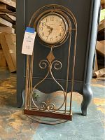 Full view of the decorative metal wall clock showing the round clock face and metal scrollwork frame, placed on the floor against a blue cabinet.