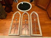 Top-down view of three arched wood and metal wall art panels on a wood floor, showing distressed white frames and metal scrollwork.