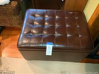 Brown padded storage ottoman with tufted cushioned top and some wear visible on surface.