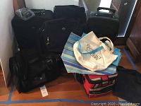Photo showing two black wheeled suitcases, multiple black soft travel bags, a red and black insulated lunch cooler bag, and several tote bags with logos.