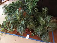 Photo showing multiple artificial plants with varied leaf textures and shapes arranged on the floor.