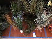 Photo showing multiple artificial plants grouped on floor with various types of grasses, floral stems, and leafy plants.