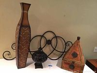 Wide shot showing four Indonesian decorative pieces including the tall vase, metal wall art, square metal container, and triangular wooden item