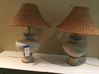 Two ceramic lamps with wicker lampshades sitting on a white surface against a beige wall. Both lamps are about 30 inches tall. One lamp has a chipped finial.