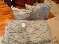 Gray pleated bedding items arranged on hardwood floor, showing two Euro shams and one pillow insert with tag