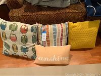 Four outdoor decorative pillows arranged on a wood floor in front of a couch. Featured are an owl print pillow, a geometric pattern pillow, a mustard yellow textured pillow, and a peach rectangular 'sunshine' pillow.
